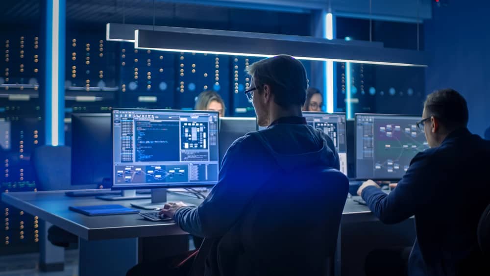 Ingenieros de ciberseguridad trabajando en un centro de datos, analizando código en múltiples monitores. La imagen transmite un ambiente de alta tecnología y concentración.