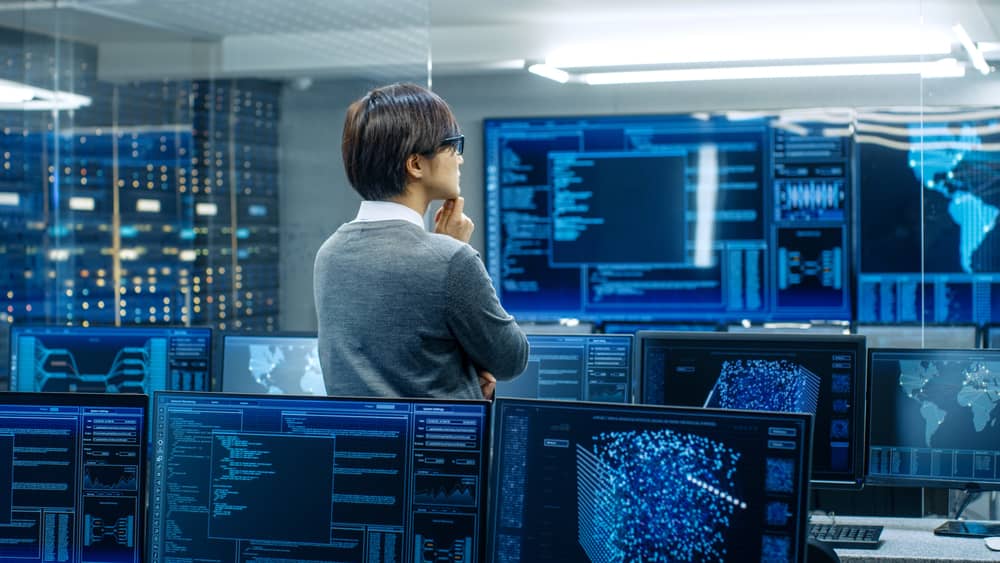 Un hombre de espaldas observa múltiples pantallas de computadoras que muestran datos y código, en un centro de control de ciberseguridad.