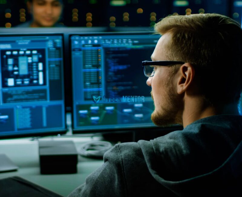 Un ingeniero de ciberseguridad con gafas se concentra en dos monitores que muestran código y datos. La imagen sugiere un entorno de trabajo de alta tecnología.