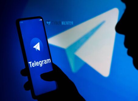 Una mano sostiene un teléfono inteligente que muestra el logo de Telegram en su pantalla. Al fondo, se ve una silueta de una persona y una imagen borrosa del logo de Telegram.