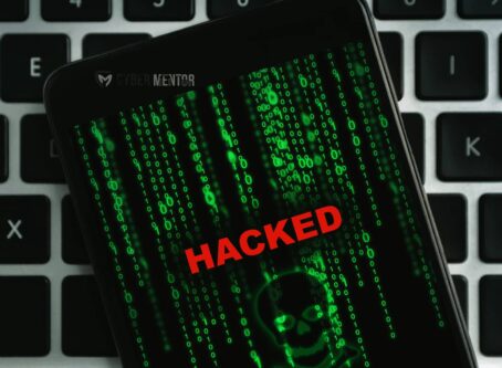 Un teléfono móvil con la palabra "HACKED" en rojo y código binario verde descendiendo sobre la pantalla, colocado sobre un teclado de computadora, simbolizando un ciberataque móvil.