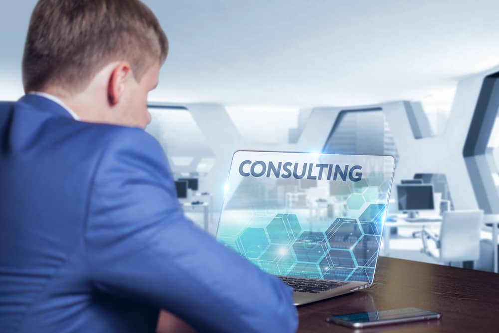 Un hombre de negocios de espaldas mirando un ordenador portátil con la palabra "CONSULTING" (consultoría) en la pantalla, en un entorno de oficina moderno.
