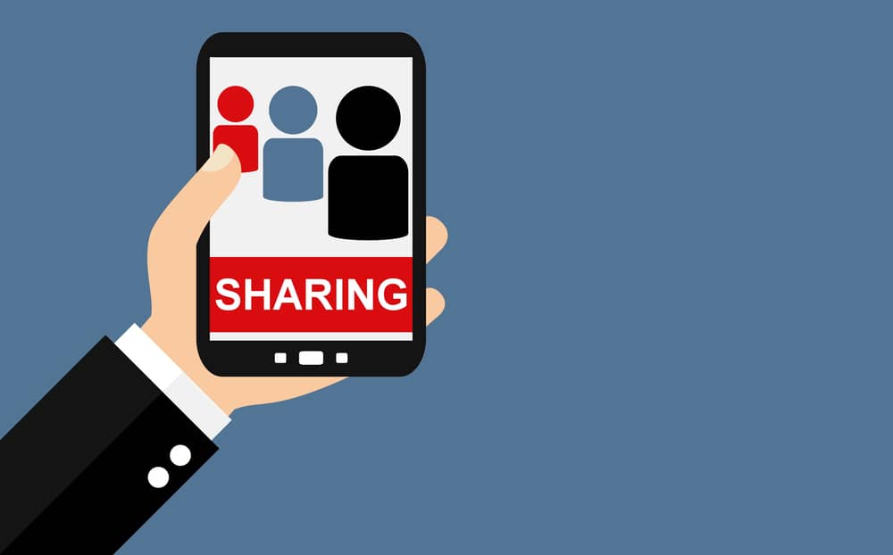 Una mano sostiene un teléfono inteligente que muestra una pantalla con iconos de personas y la palabra "SHARING" (Compartir), ilustrando el concepto de compartir información de forma segura.