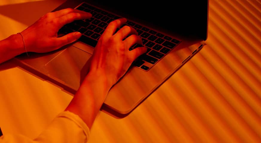 Manos de una persona escribiendo en un teclado de ordenador portátil, con una iluminación naranja intensa, simbolizando la acción preventiva o la conciencia en ciberseguridad.