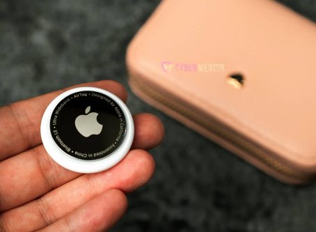 Mano sosteniendo la parte trasera de un AirTag de Apple, mostrando el logo y detalles técnicos, con una cartera rosa al fondo.