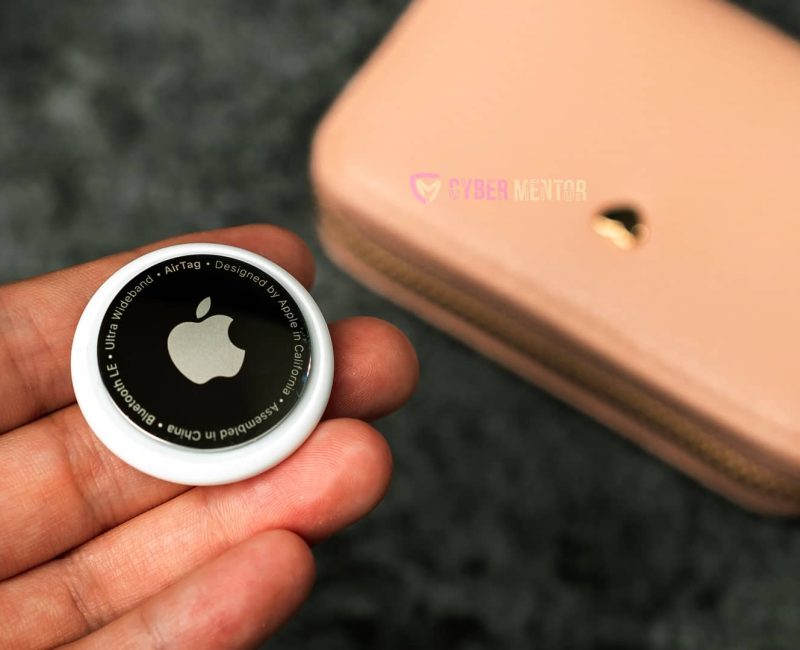 Mano sosteniendo la parte trasera de un AirTag de Apple, mostrando el logo y detalles técnicos, con una cartera rosa al fondo.